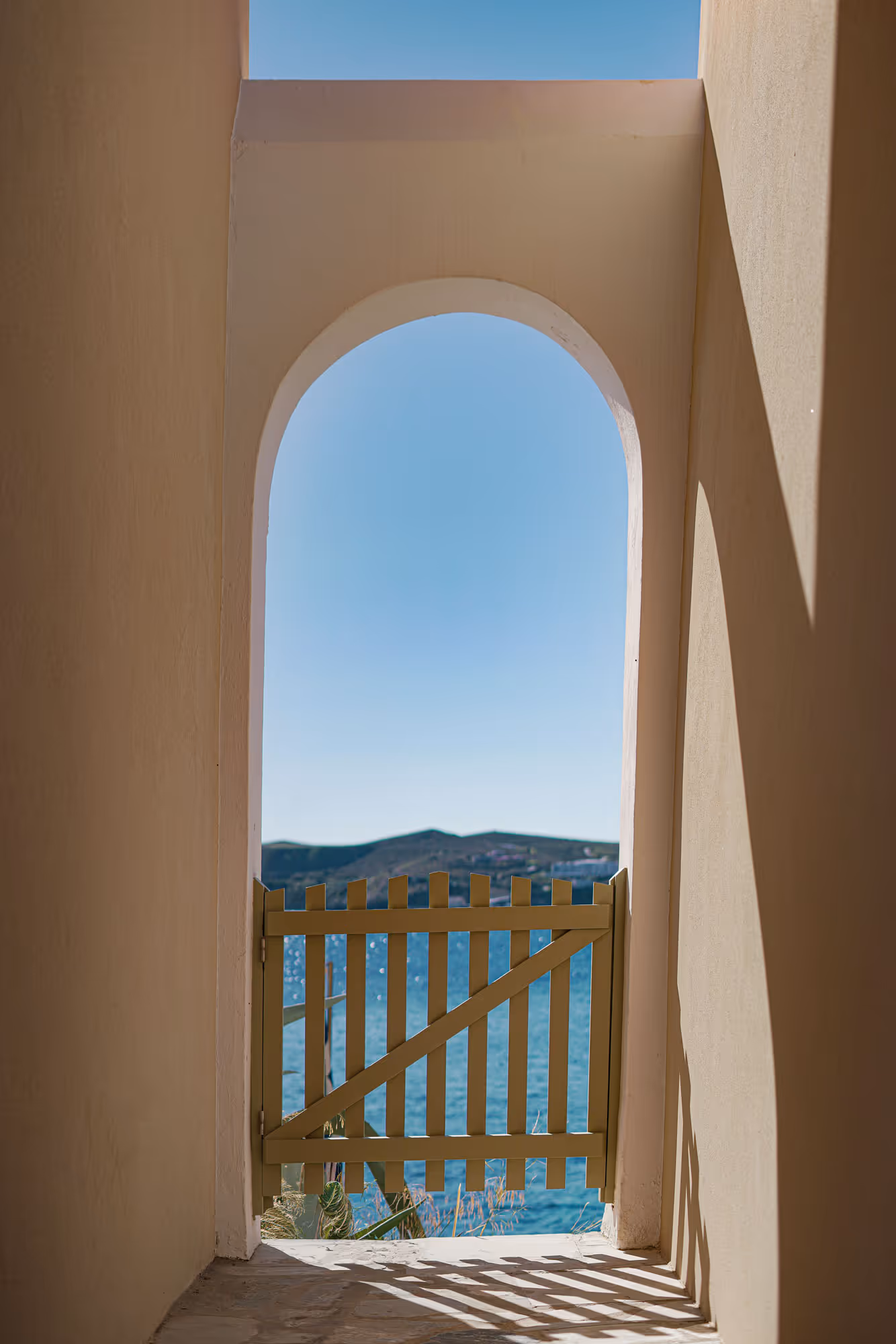 Sunlit archway with a wooden picket gate overlooking a blue sea and distant hills.
