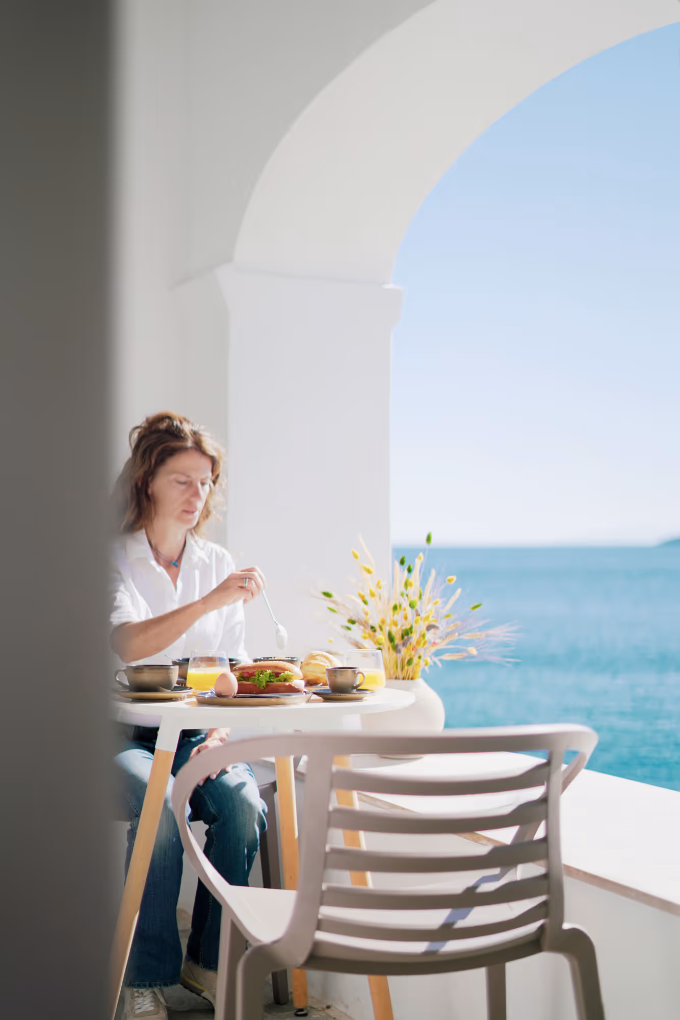 Woman eating breakfast at a white table on a terrace overlooking the sea under an archway.